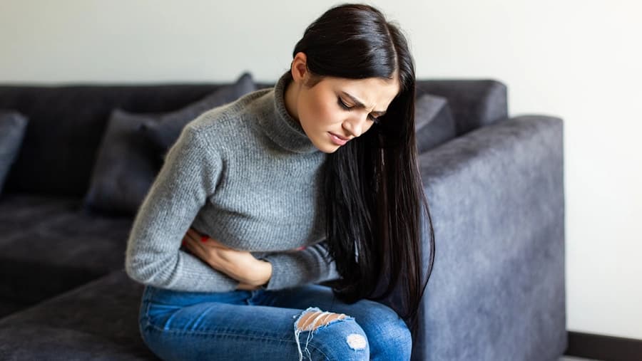 Mujer joven con cabello oscuro sostiene su abdomen con gesto de dolor sentada en un sofá gris.