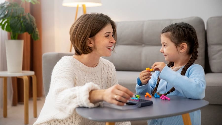 Terapeuta sonriente jugando con piezas de colores junto a una niña con trenzas en mesa redonda gris.